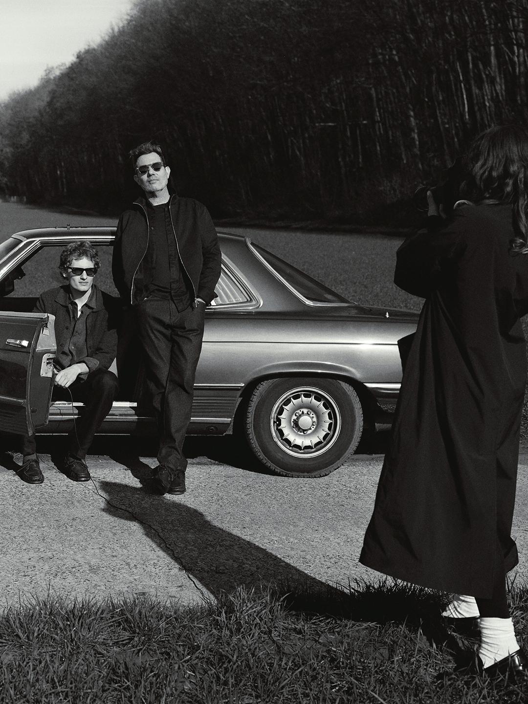 Imagem a preto e branco de duas pessoas com óculos de sol junto a um carro clássico, enquanto uma terceira pessoa as fotografa numa estrada.