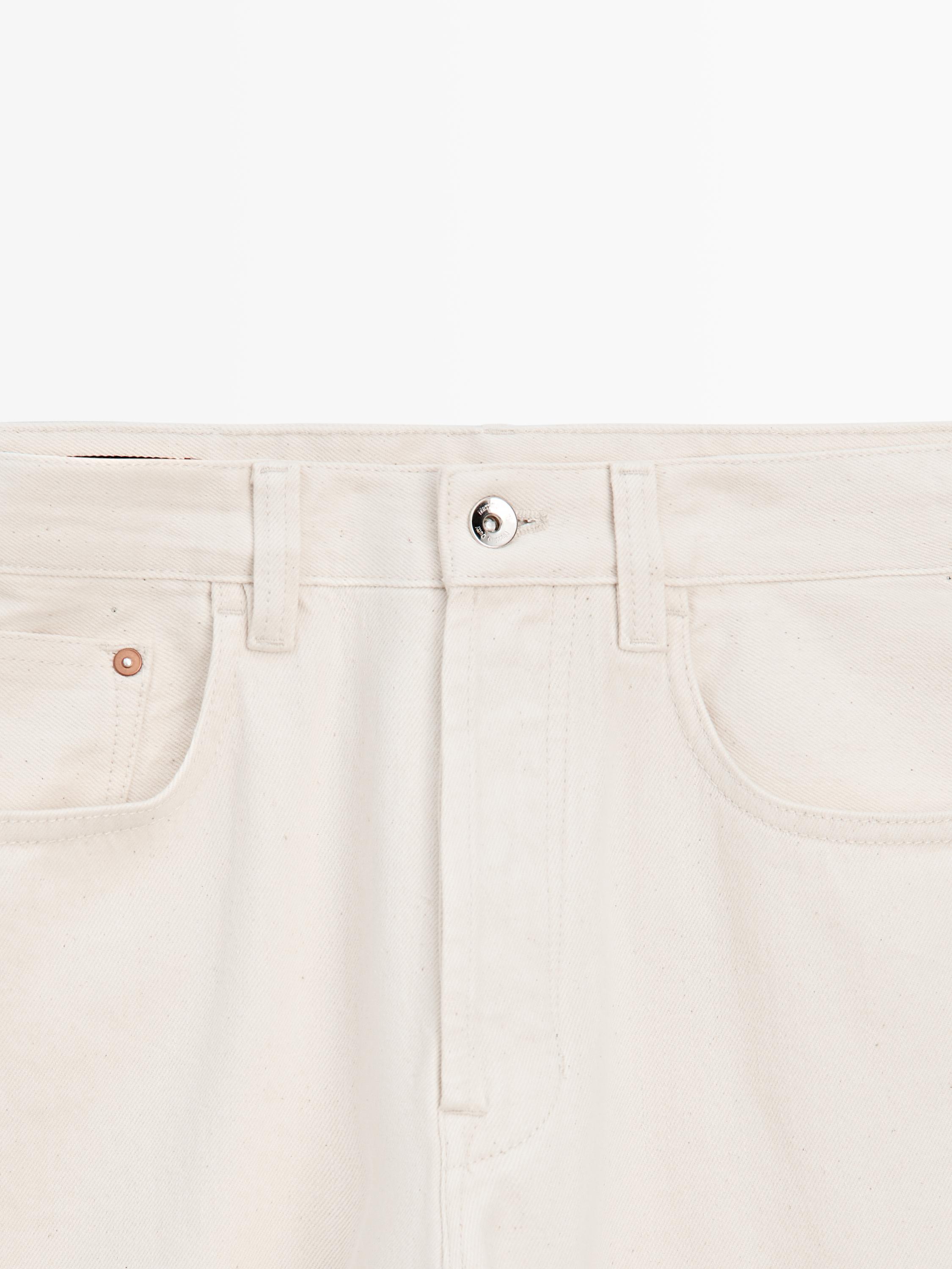 Medium shot of the waist of raw denim jeans with a metal button and visible front pockets.