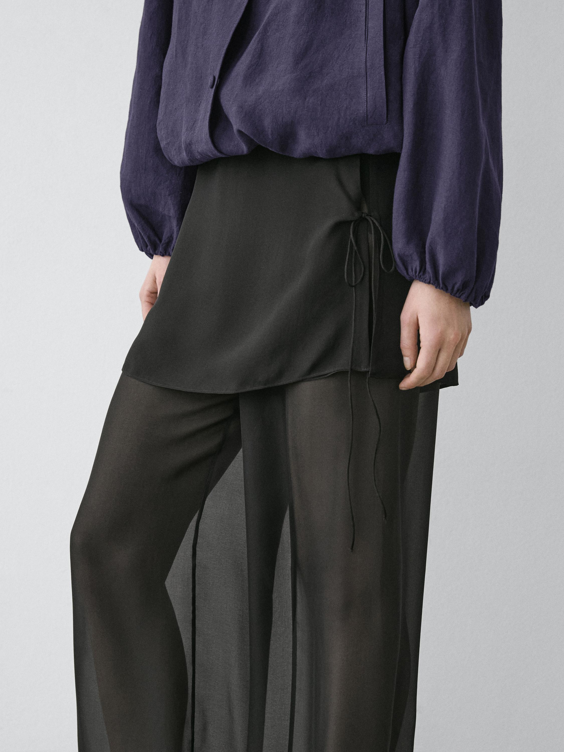 Plano médio de calças pretas fluídas com camada superior curta e laço lateral, combinado com blusa roxa de mangas compridas.