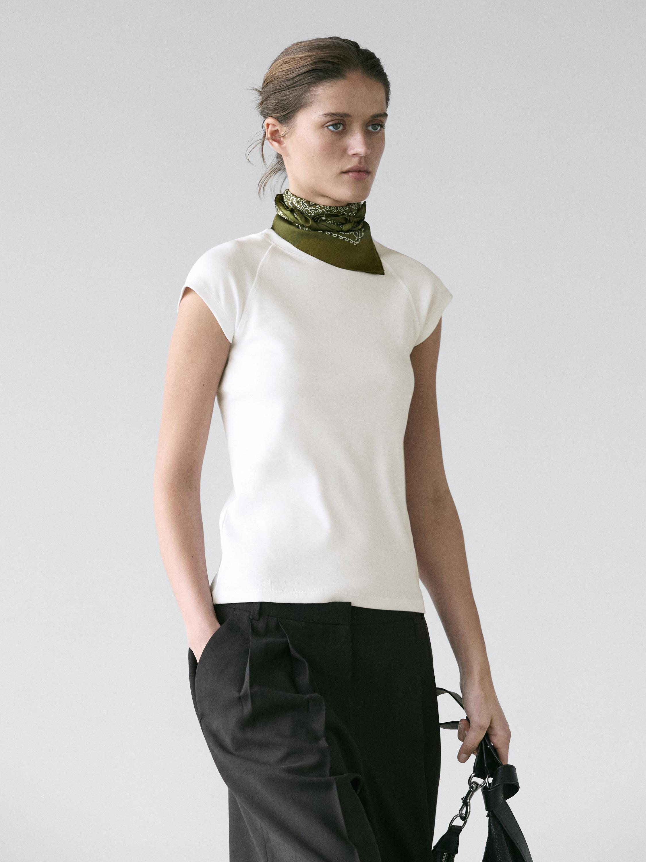 Ensemble composé d'un t-shirt blanc à manches courtes et d'un pantalon noir à plis avec des poches latérales. Foulard vert autour du cou.