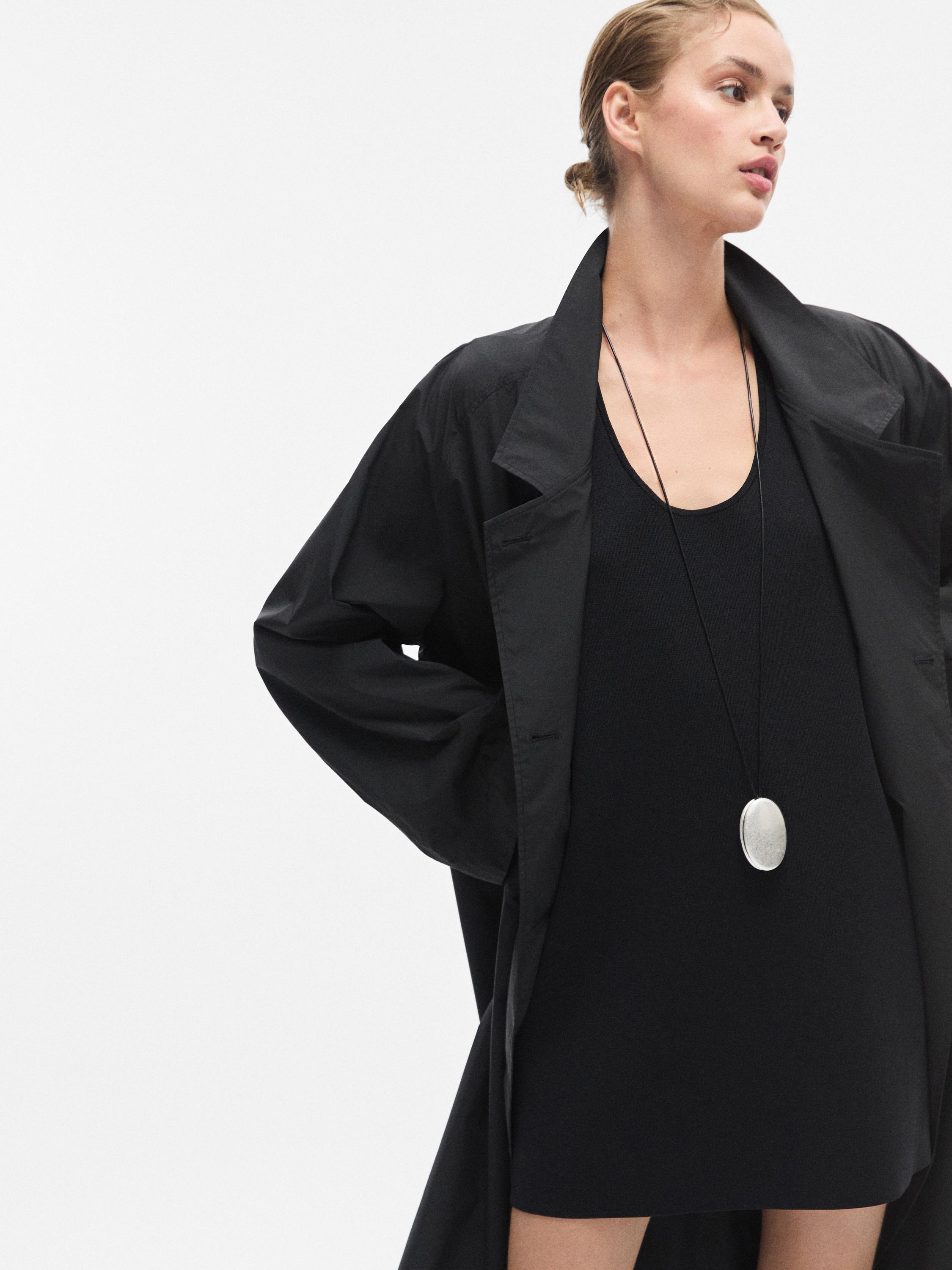 Μεσαίο πλάνο ενός μαύρου, ρέοντος μίνι φορέματος με στρογγυλή λαιμόκοψη, αμάνικο, σε συνδυασμό με ένα φαρδύ μαύρο παλτό.