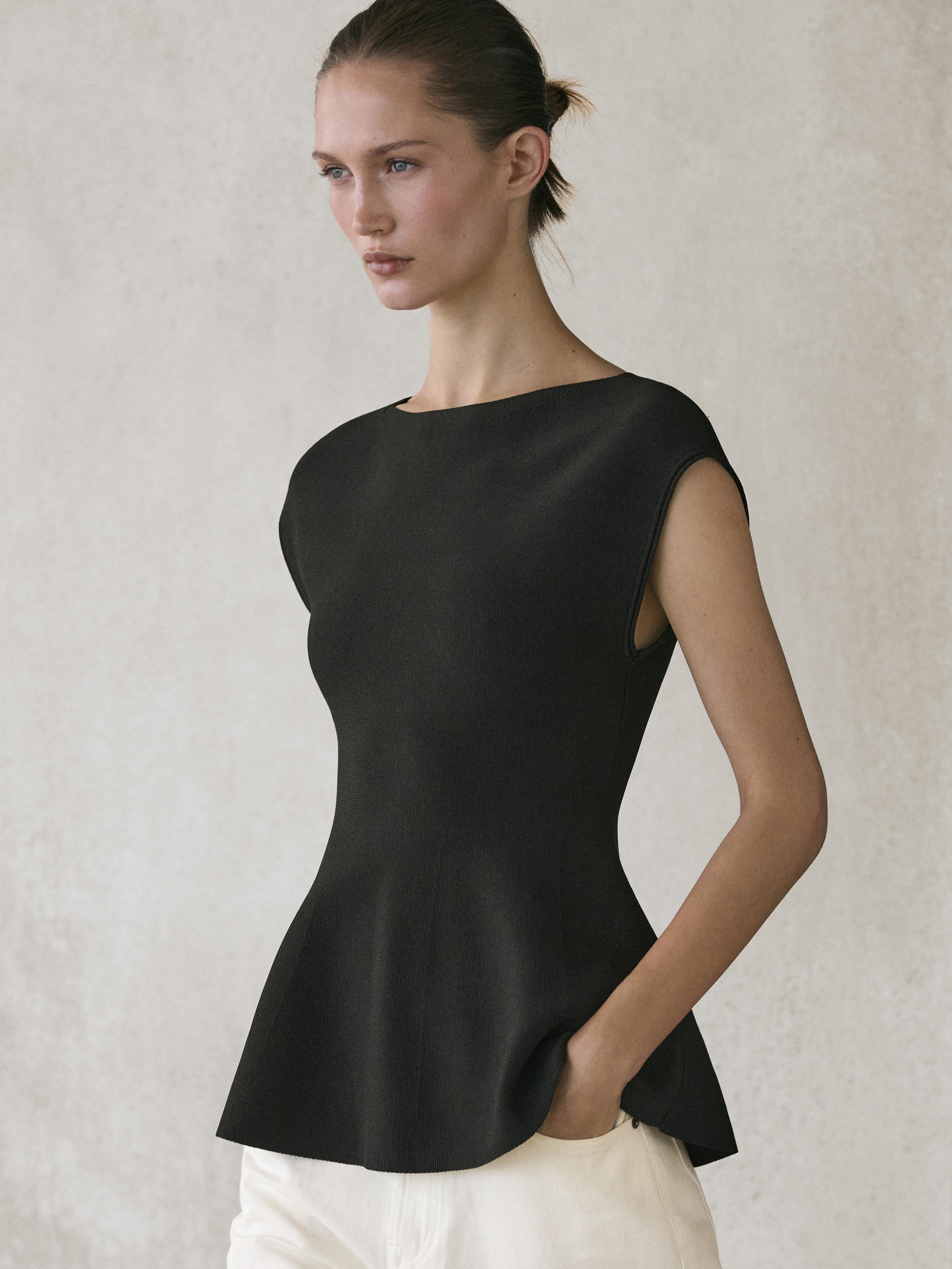 Plan moyen d'un top noir en maille avec col rond et coupe péplum, sans manches, assorti d'un pantalon blanc.