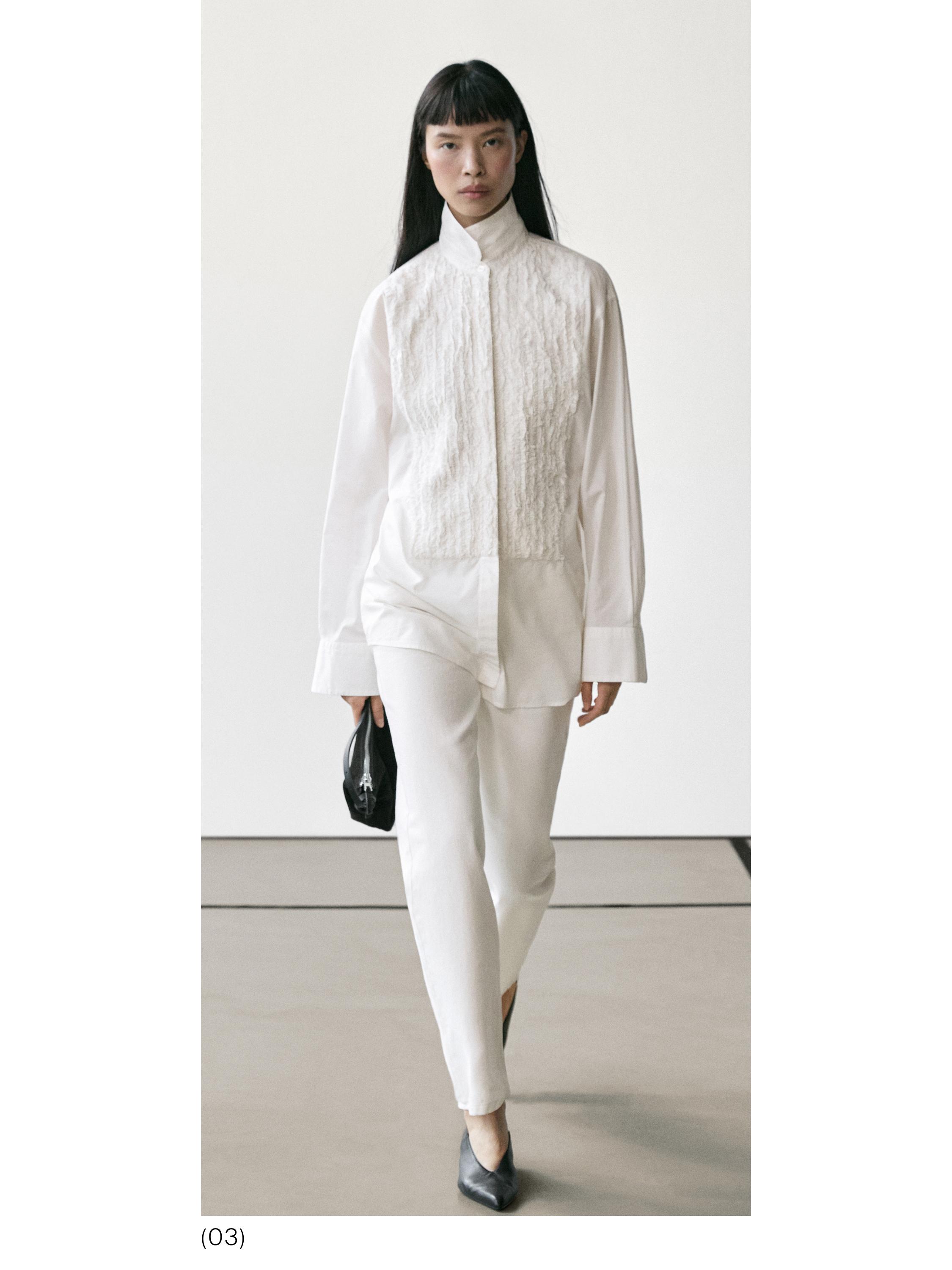 Komplet me këmishë të bardhë me panel me teksturë dhe pantallona të bardha. Mban çantë të zezë dhe taka të zeza.