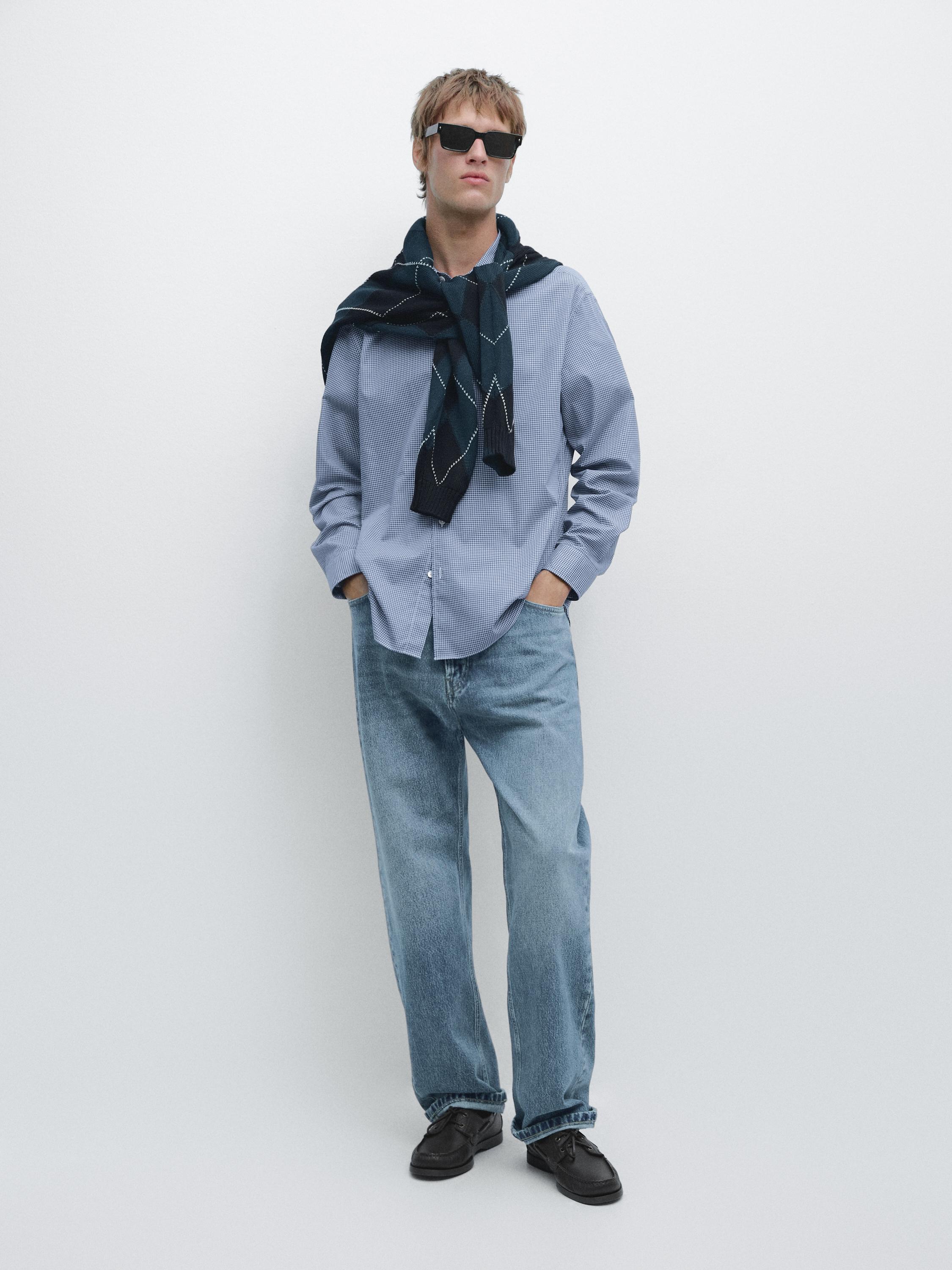 Blue long-sleeved shirt, blue jeans, and black shoes set. Dark sweater tied around the neck.