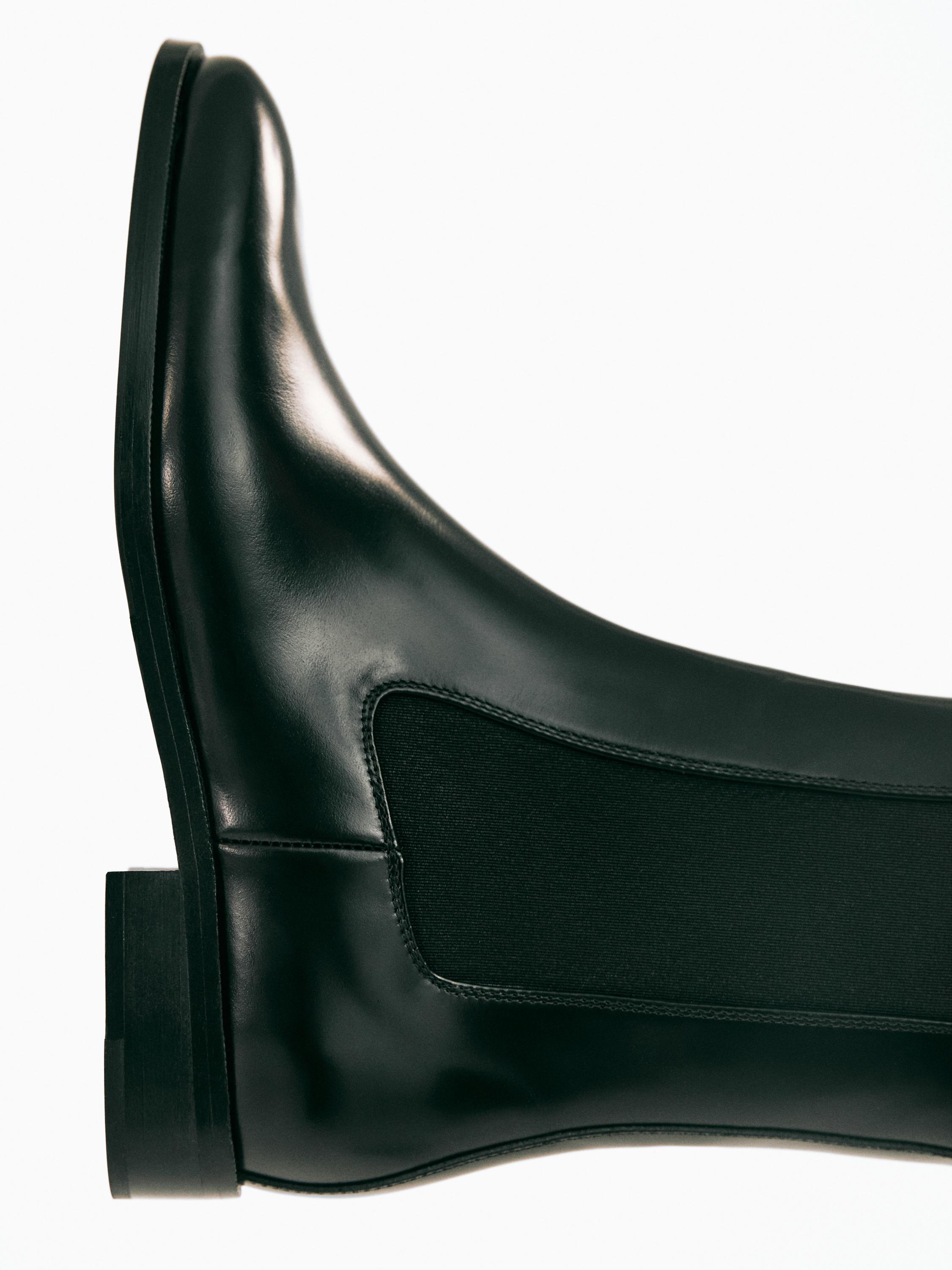 "Détail de botte noire à la cheville avec empiècement élastique latéral et semelle plate."