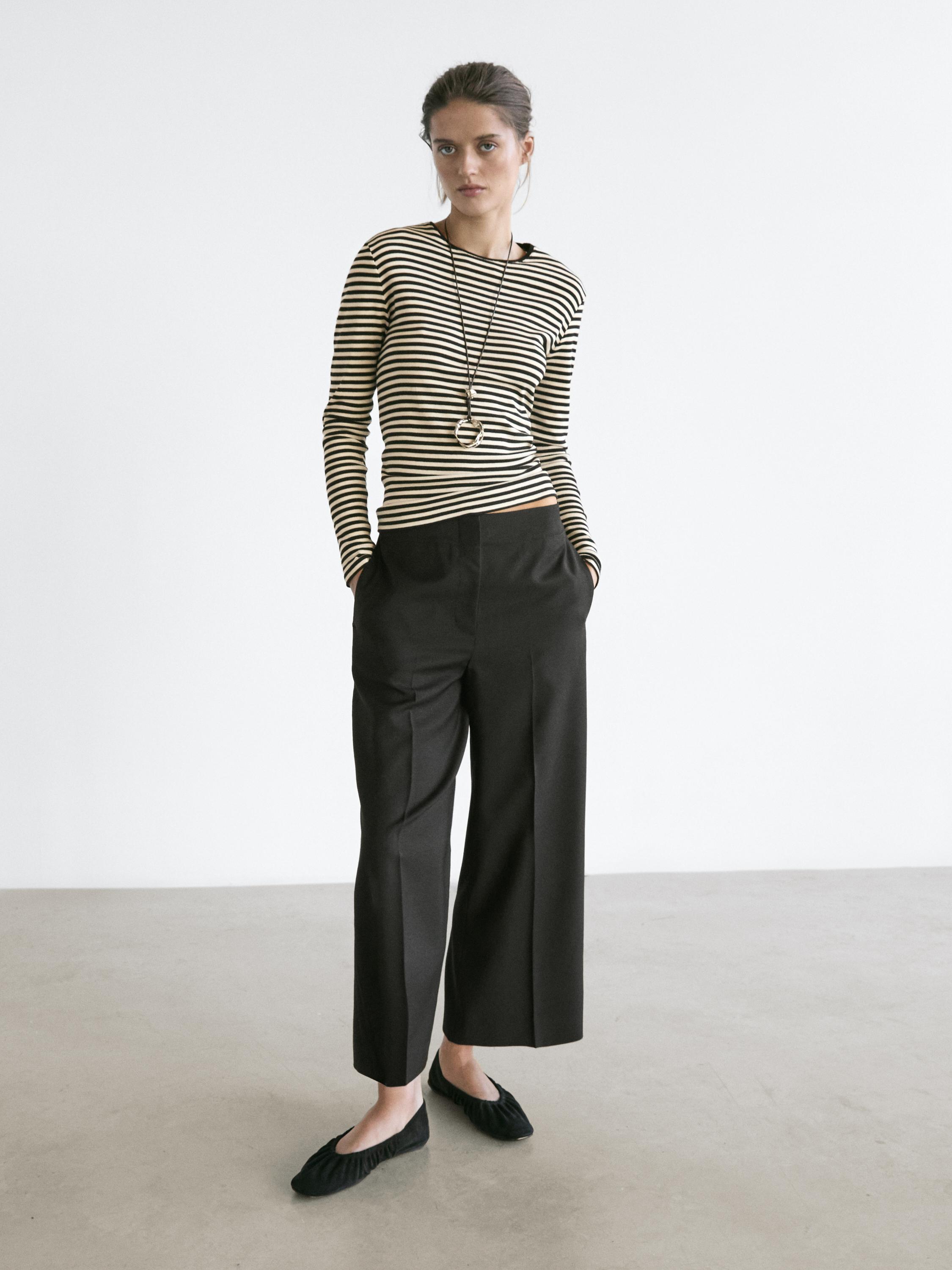 Komplet me pantallona të zeza të gjera crop me xhepa anësorë dhe bluzë me mëngë të gjata me vija bardh e zi.