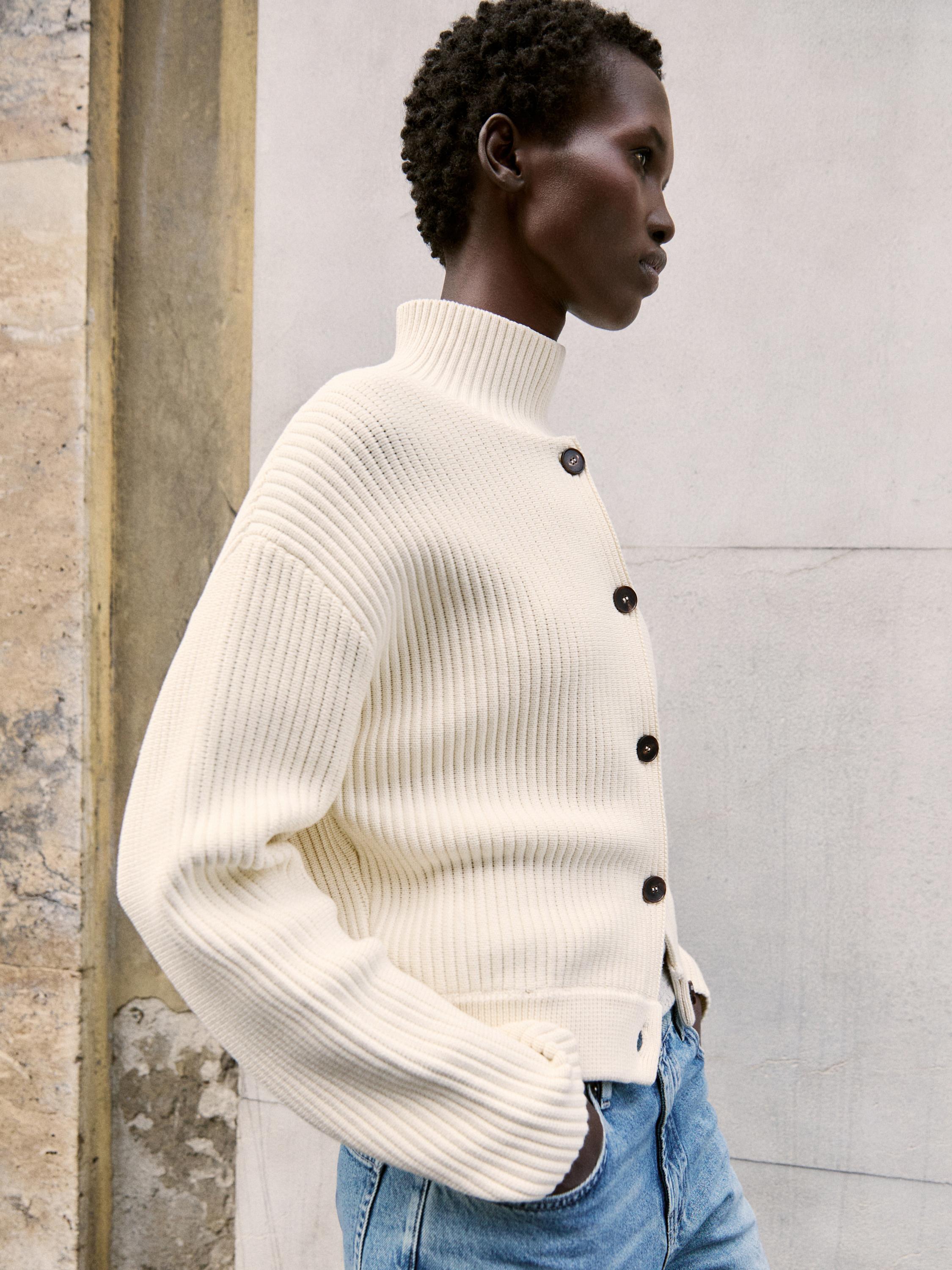 Plan moyen d'un pull en maille épaisse blanc à col roulé et boutons foncés, associé à un jean bleu.