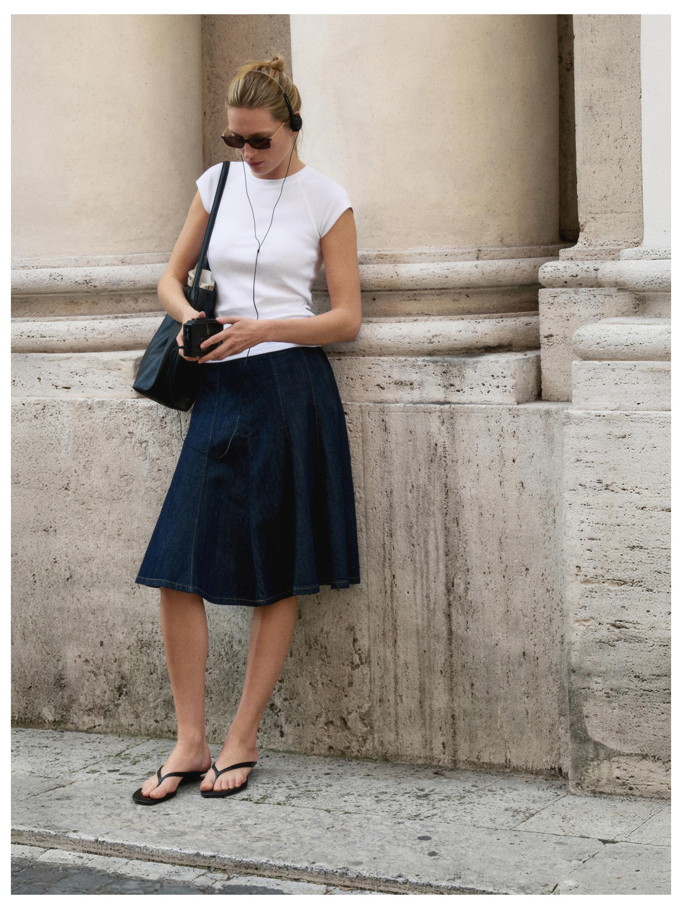 Beyaz kolsuz tişört, lacivert diz boyu etek ve siyah sandalet kombini. Siyah çanta taşıyor.