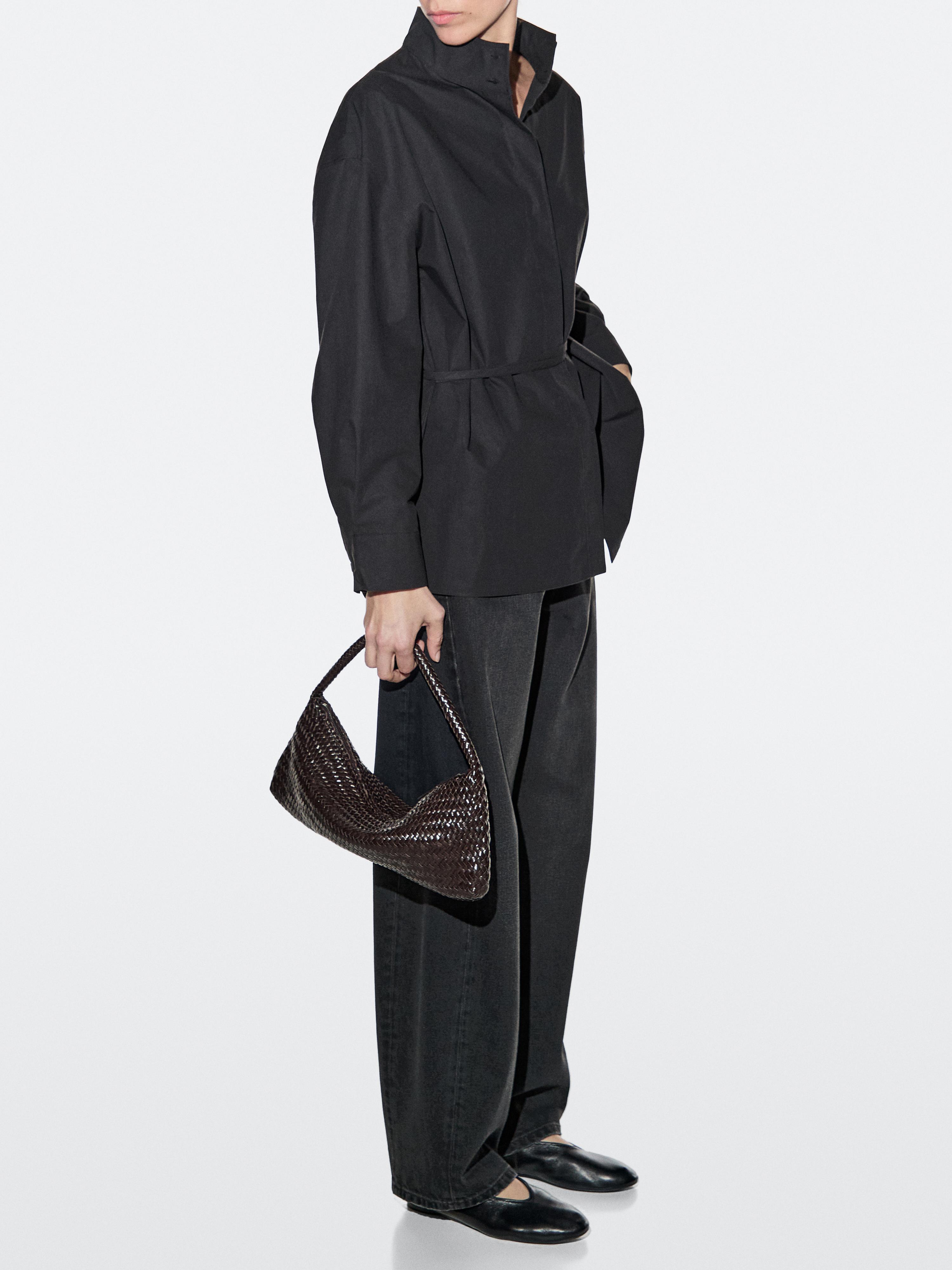 Conjunto de casaco preto com cinto, calças escuras e mala castanha entrançada.