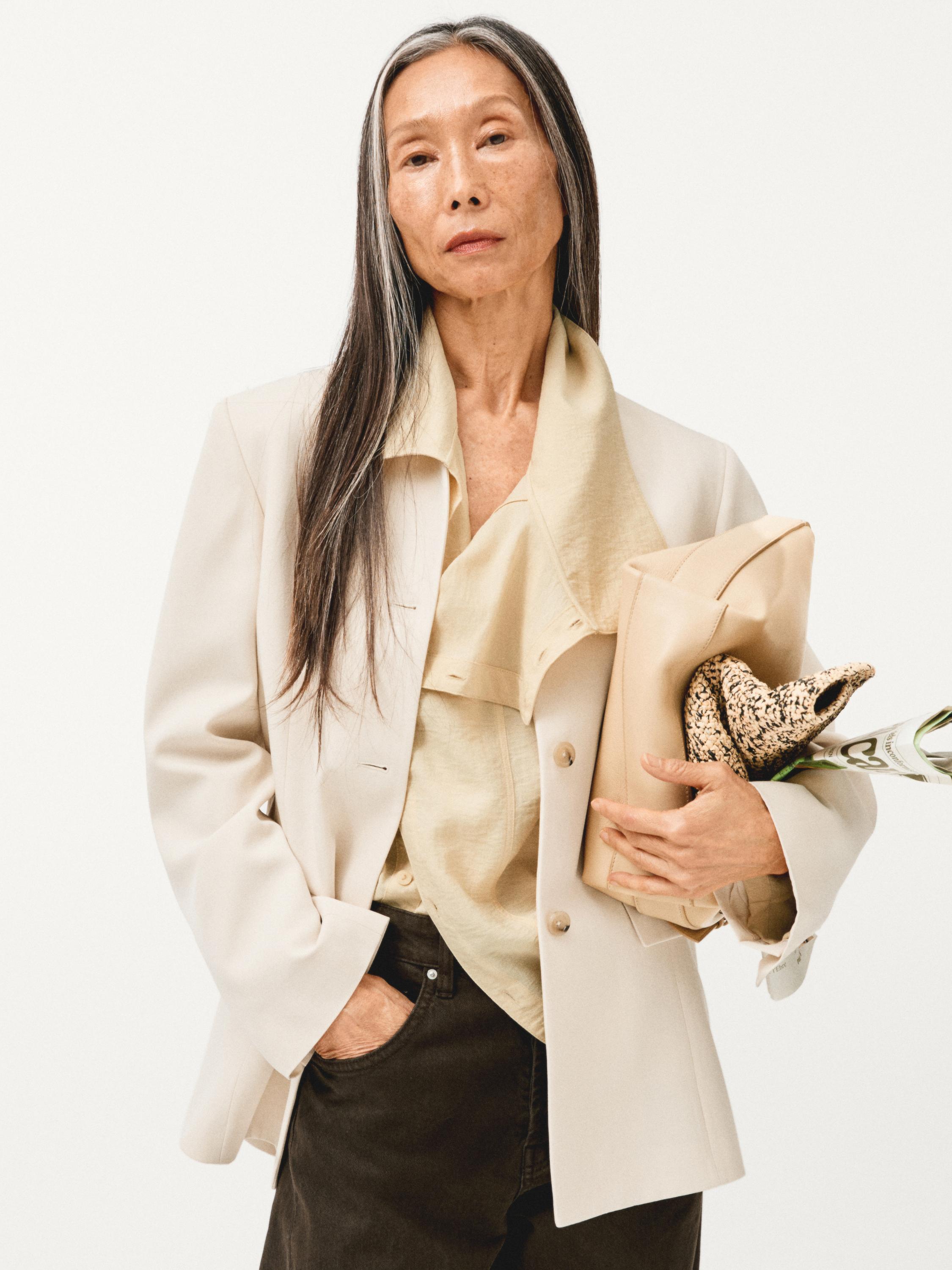 Smėlio spalvos švarko, kreminės palaidinės ir tamsių kelnių komplektas. Švarkas su sagomis ir matoma šonine kišene.