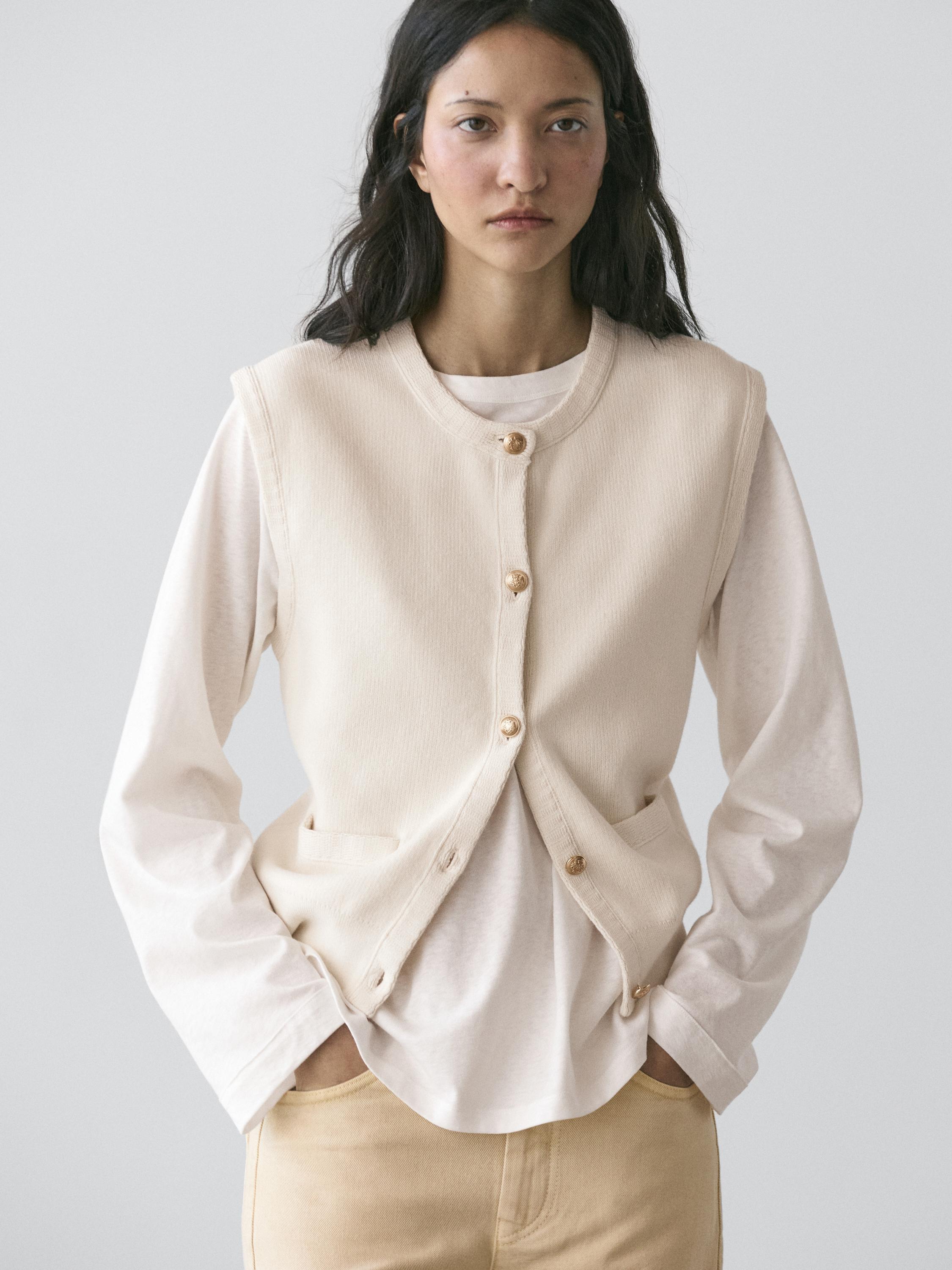 plaintext Botoiak eta alboetako poltsikoak dituen txaleko gordinaren plano ertaina, mahuka luzeko kamiseta zuri baten gainean.