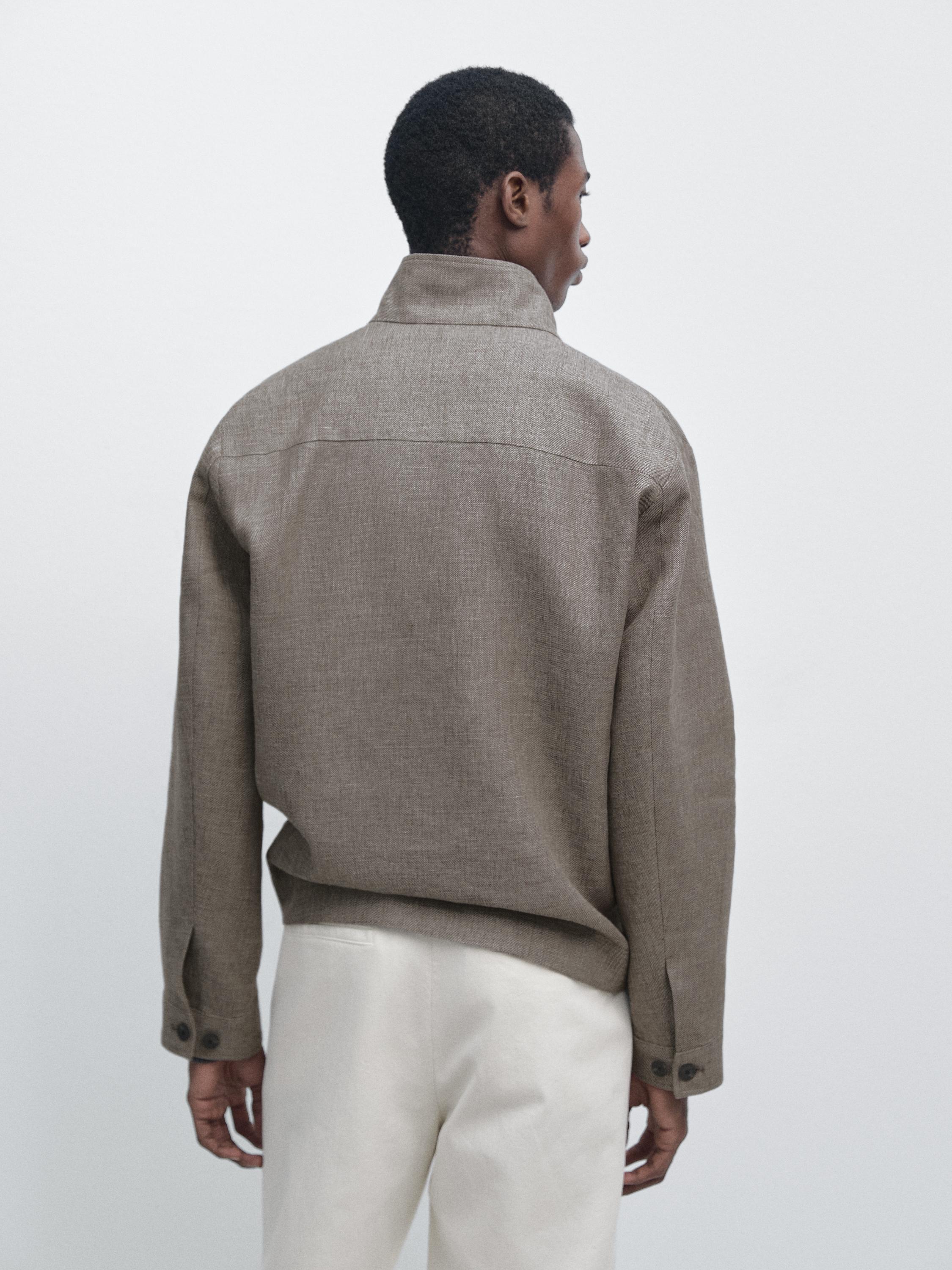 Комплект із бежевої сорочки-жакета, вигляд ззаду, з високим коміром та манжетами на ґудзиках, у поєднанні з білими штанами.