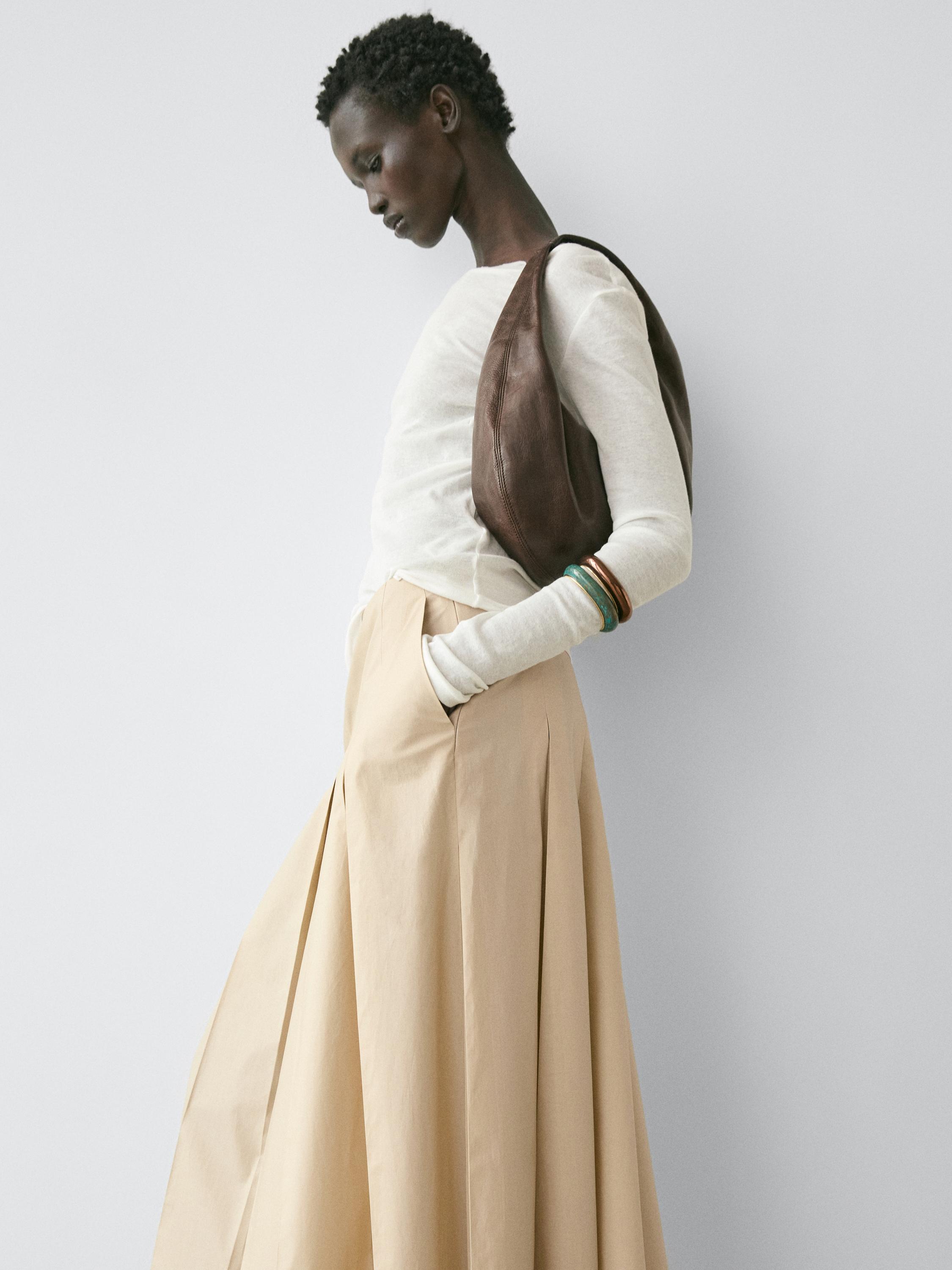 Ensemble composé d'un t-shirt blanc semi-transparent à manches longues et d'une jupe plissée beige, avec un sac marron porté à l'épaule.