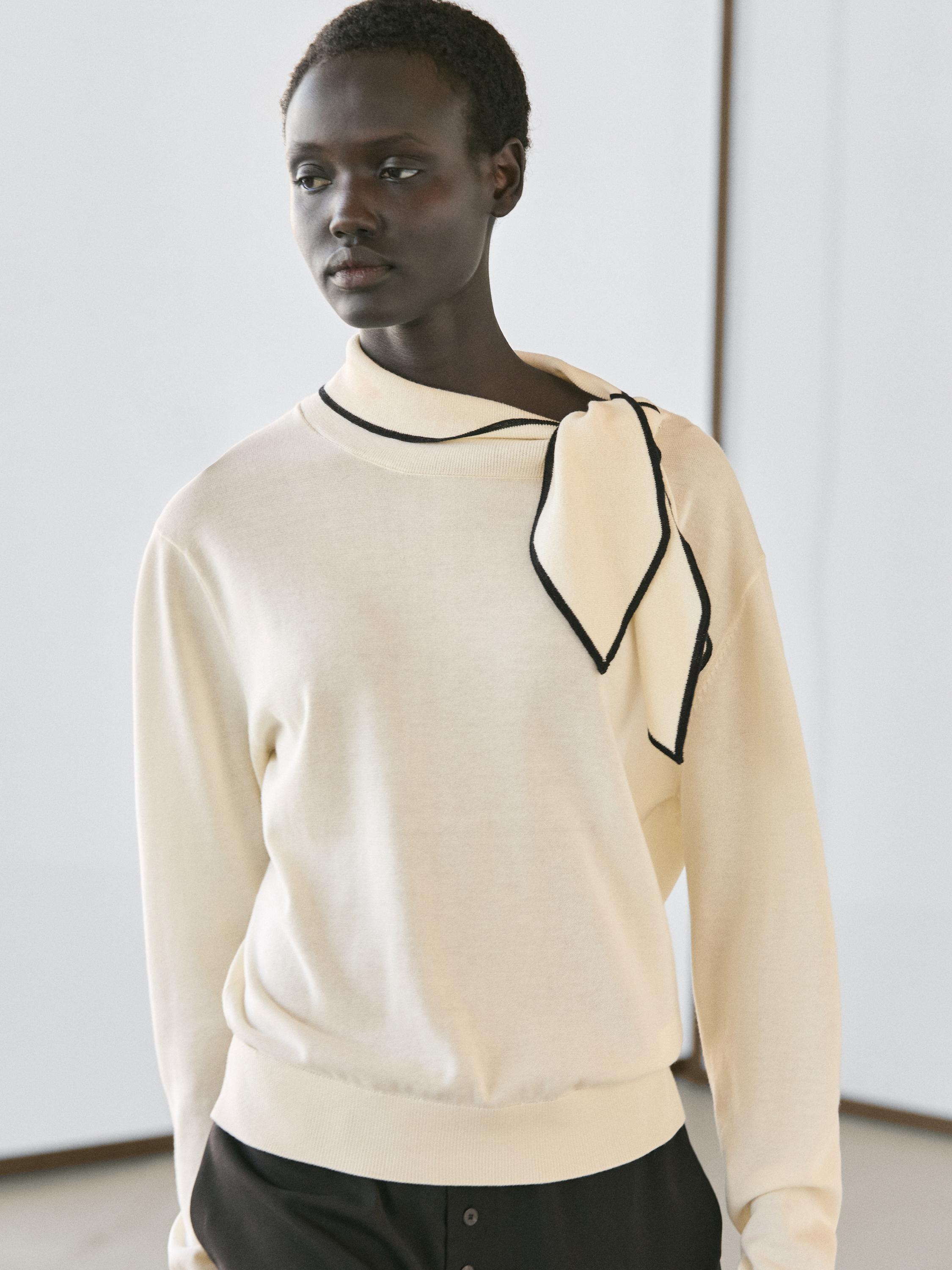 Plan moyen d'un pull écru avec un détail de foulard noir au col, des manches longues et une coupe ample.