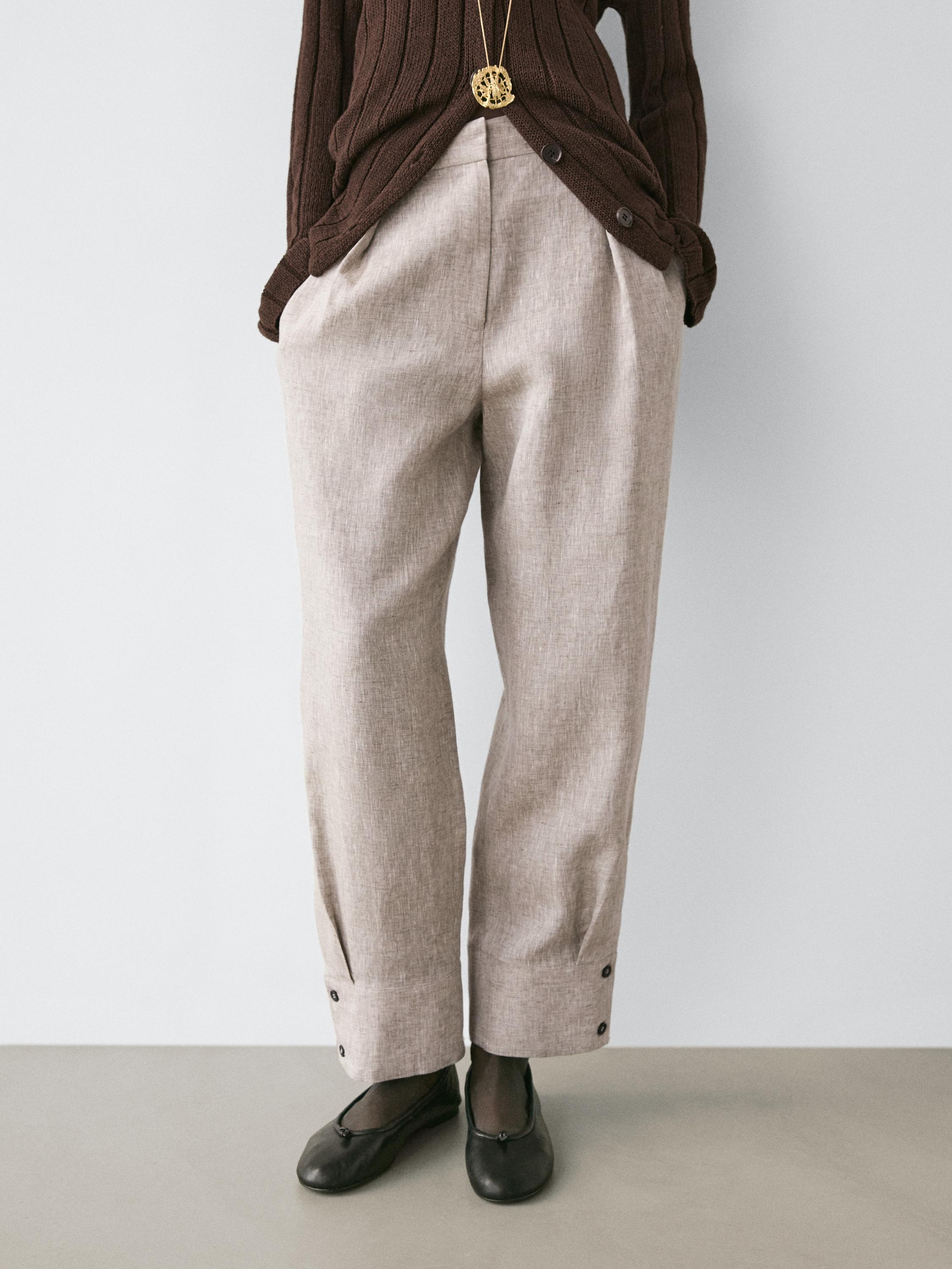 طقم بنطلون بيج بتفاصيل إبزيم وأزرار عند الحواف، وكنزة بنية وحذاء أسود.