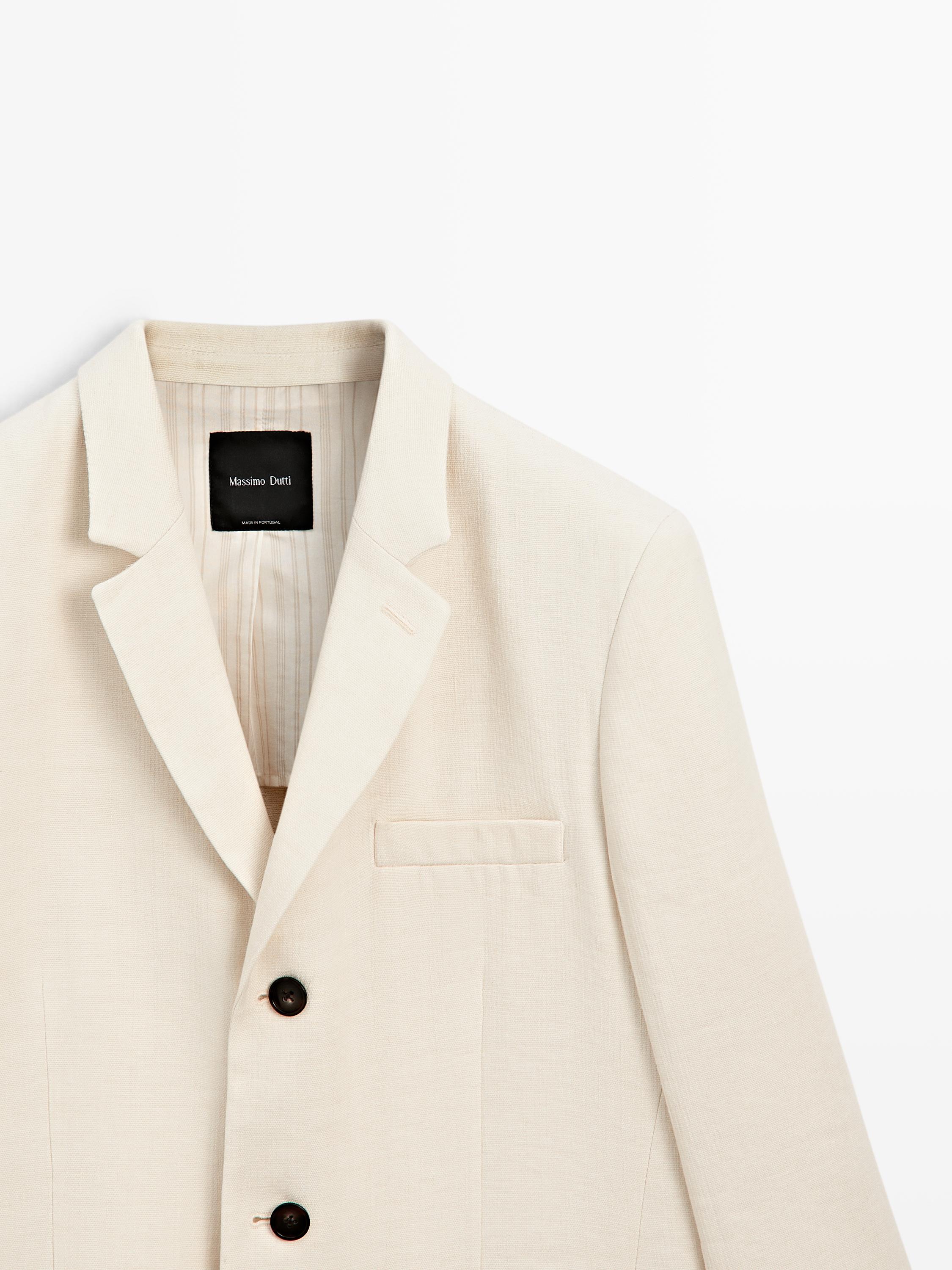 Halbaufnahme eines cremefarbenen Blazers mit klassischem Revers, zwei dunklen Knöpfen und einer aufgesetzten Tasche ohne Patte. Sichtbares Etikett innen.