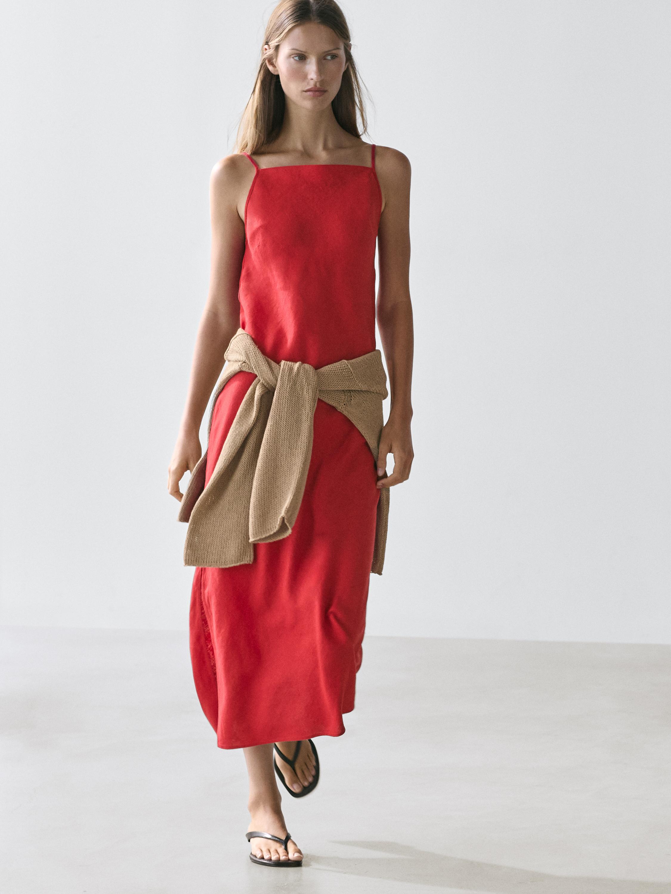 Ensemble robe midi rouge à décolleté droit et fines bretelles, avec pull beige noué à la taille et sandales noires.