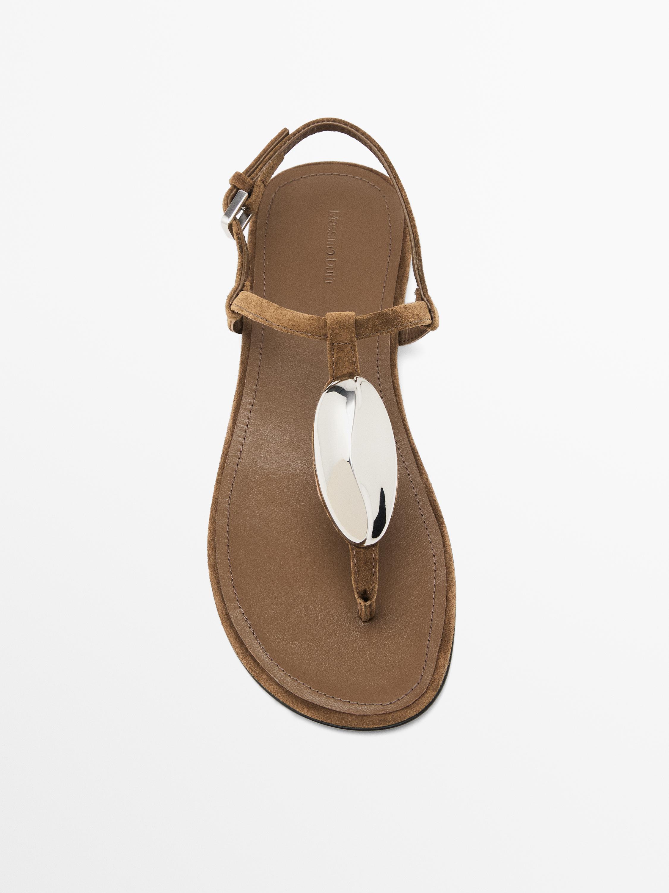 Front view of a brown sandal with an oval metallic detail on the upper and an adjustable strap.