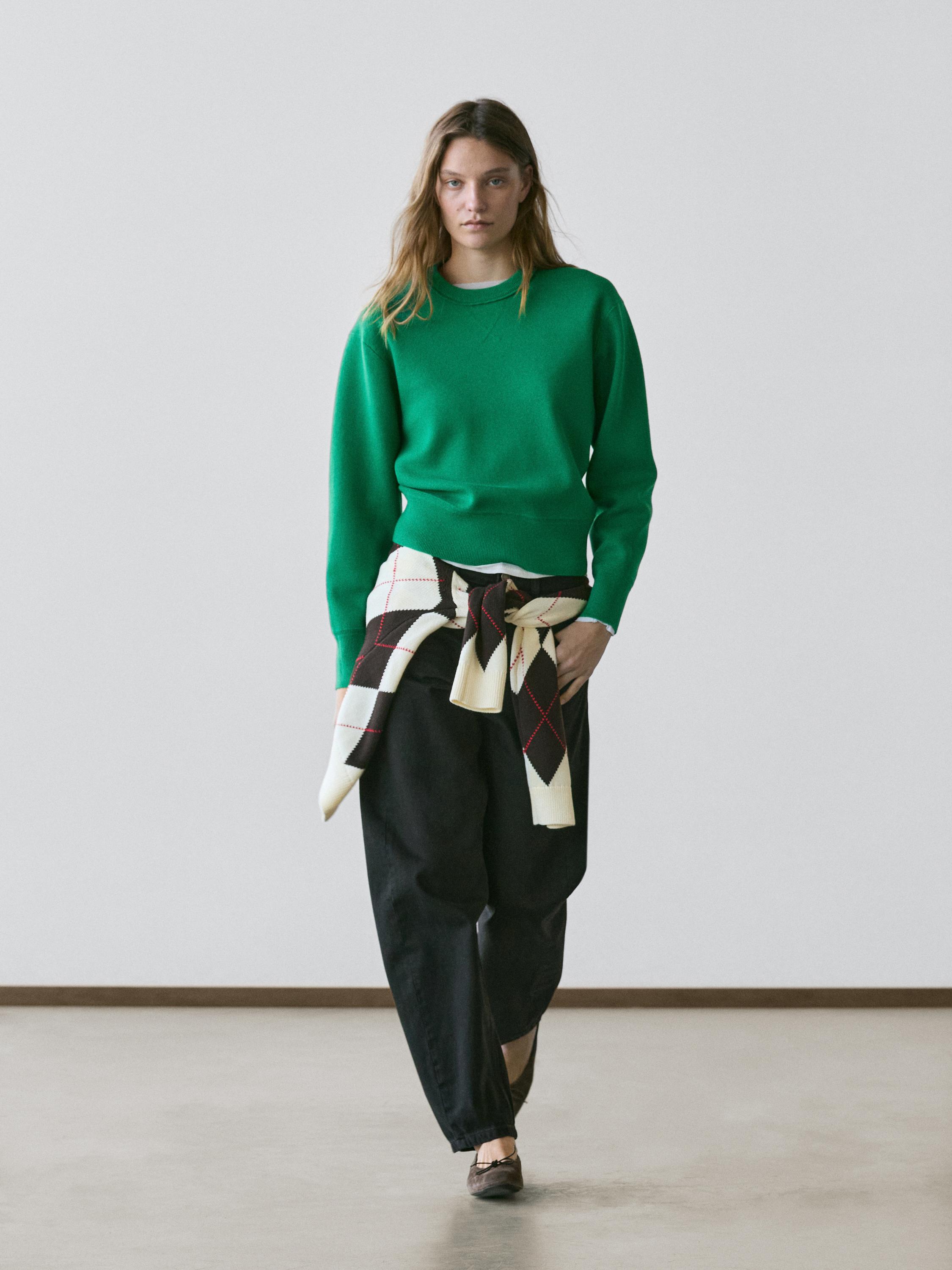 Set aus billardgrünem Pullover, weiter schwarzer Hose und einem an der Taille gebundenen Schal mit Rautenmuster.